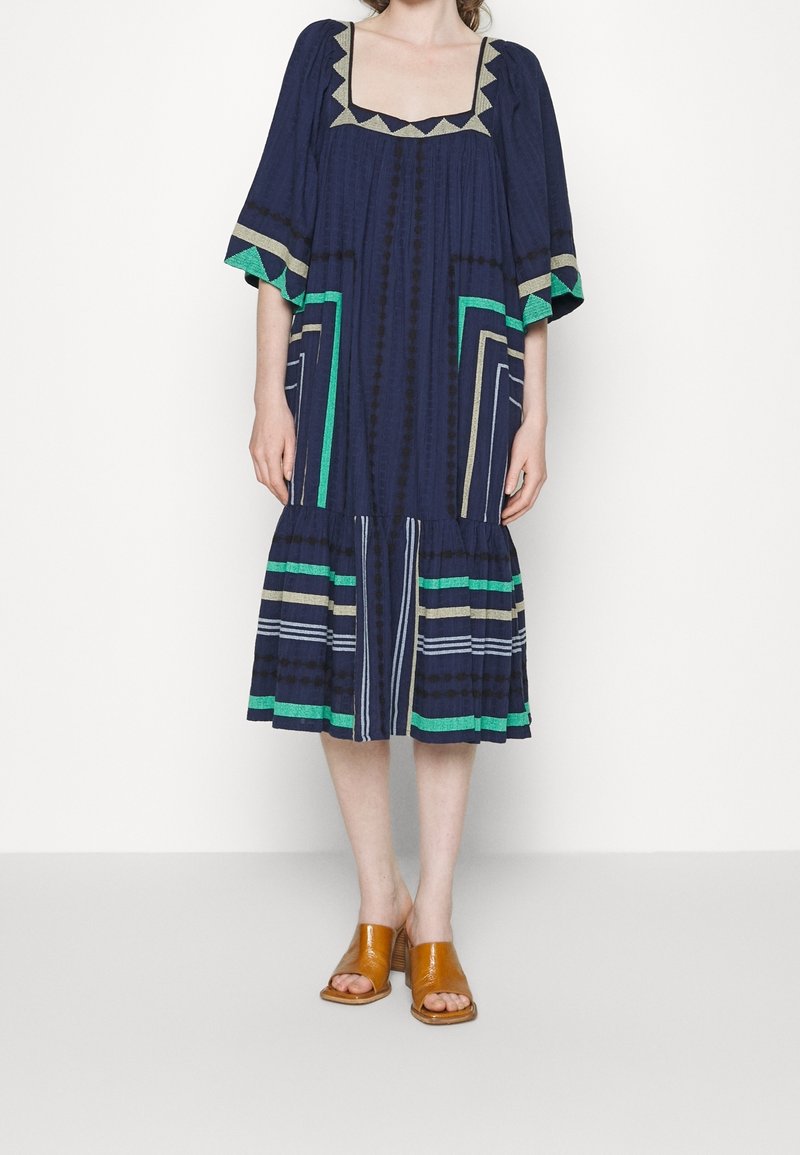 Femme portant une robe midi bleu marine avec des motifs géométriques verts, beiges et blancs, associée à des sandales à talons blocs ouvertes à l'avant couleur tan.