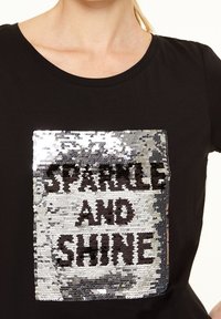 Camiseta negra con un gráfico rectangular de lentejuelas que presenta el texto "BRILLA Y RESPLandece" en letras negras sobre un fondo plateado.