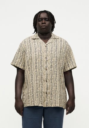 Mand med mørk hud og dreadlocks iført en beige kortærmet skjorte med små bladmønstre og mørkeblå bukser.