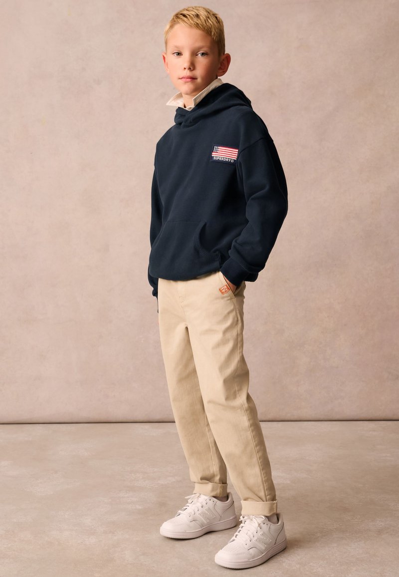 Felpa navy con tasca frontale e logo, pantaloni beige a sigaretta e sneakers bianche. Tessuto morbido, vestibilità comoda, design casual.