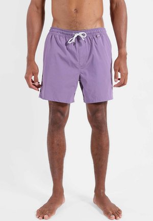 Homme portant un short de bain violet clair jusqu'à mi-cuisse avec un cordon blanc, debout pieds nus sur un fond uni.