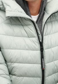 Chaqueta acolchada verde claro con diseño acolchado horizontal, cremallera frontal y capucha. La capa interior presenta tela gris con un cuello alto.