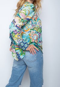 Kvinde i farverig, mønstret bluse med lange ærmer og lyseblå denimjeans, hånd hvilende på hoften, mod en ensfarvet lys baggrund.