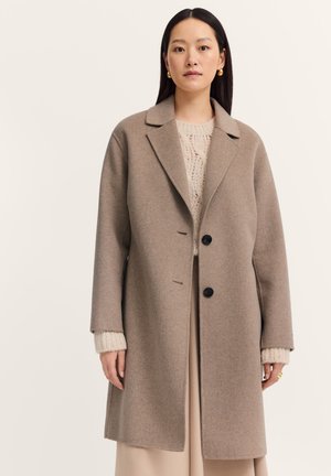 Beige Wollmischungsjacke mit einem Reverskragen, zwei schwarzen Knöpfen, frontalen Taschen und einem lockeren Schnitt. Getragen über einem leichten Strickpullover.