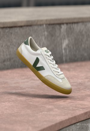 Hvid sneaker med grønne accenter, lavet af ruskind og lærredsmaterialer, rund tå, snørelukning og en sål af gummi. Har en fremtrædende side-logo.