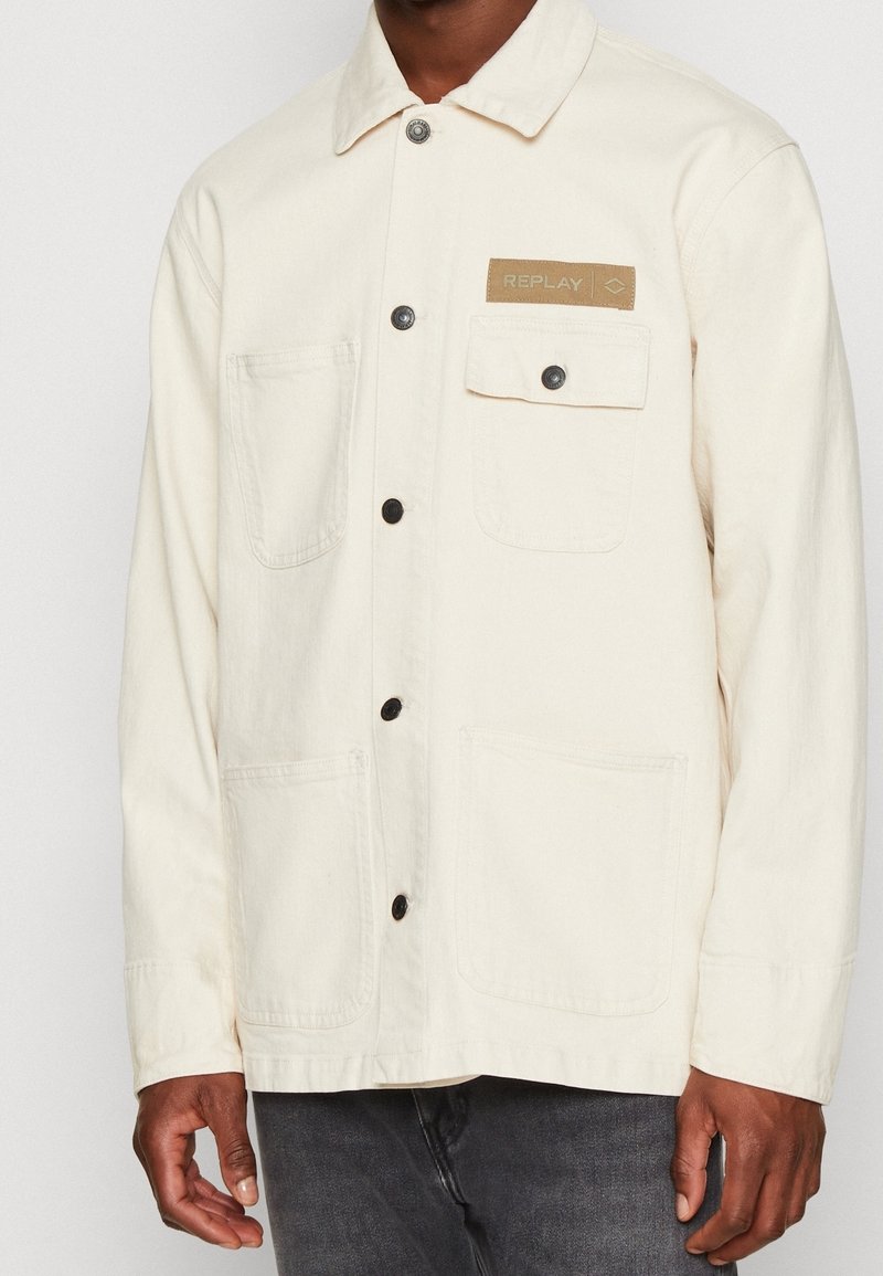 Chaqueta de color crema con cuello, botones negros, dos bolsillos frontales y un parche con el nombre de la marca "REPLAY". Tejido texturizado, diseño casual.