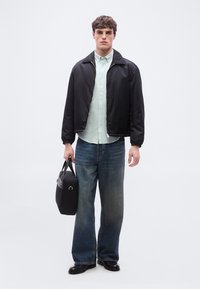 Νέος άνδρας που φοράει μαύρο μπουφάν με φερμουάρ, ανοιχτοπράσινο πουκάμισο με κουμπιά, φαρδιά μπλε τζιν παντελόνια, μαύρα παπούτσια, κρατώντας μια μαύρη τσάντα στο πλάι του.