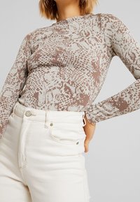 Langärmliges Oberteil in einem beigen und braunen Schlangenmuster, gefertigt aus einem eng anliegenden Stoff mit glatter Textur. Kombiniert mit hellen Jeans.