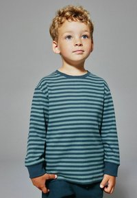 Młody chłopiec z kręconymi blond włosami i niebieskimi oczami, ubrany w niebiesko-zieloną koszulę z długim rękawem, stojący na szarym tle.