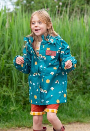 Teal raincoat with rainbow and sun patterns, featuring a chest pocket, paired with orange shorts and red boots. Outdoor setting with greenery.