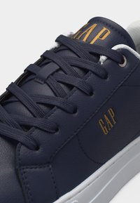 Λευκό μπλε δερμάτινο αθλητικό παπούτσι με μπλε κορδόνια, λευκή σόλα και χρυσό λογότυπο "GAP" στη γλώσσα και στην πλευρά.