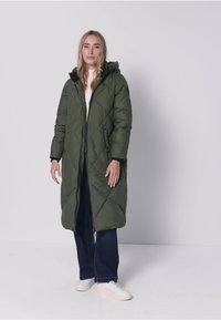 Long manteau matelassé vert avec matelassage en losanges, une capuche et des poches latérales. Porté sur un pull léger et un jean large foncé. Baskets blanches.