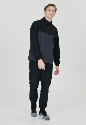 Veste légère noire et grise avec fermeture éclair à l'avant, col haut et poignets élastiques ; associée à un pantalon noir et des chaussures de sport grises.