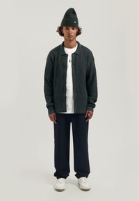 Donkergroene, gestructureerde cardigan met een kraag, knopen en een bijpassende beanie. Draagt over een wit T-shirt en gecombineerd met marineblauwe broek en witte sneakers.