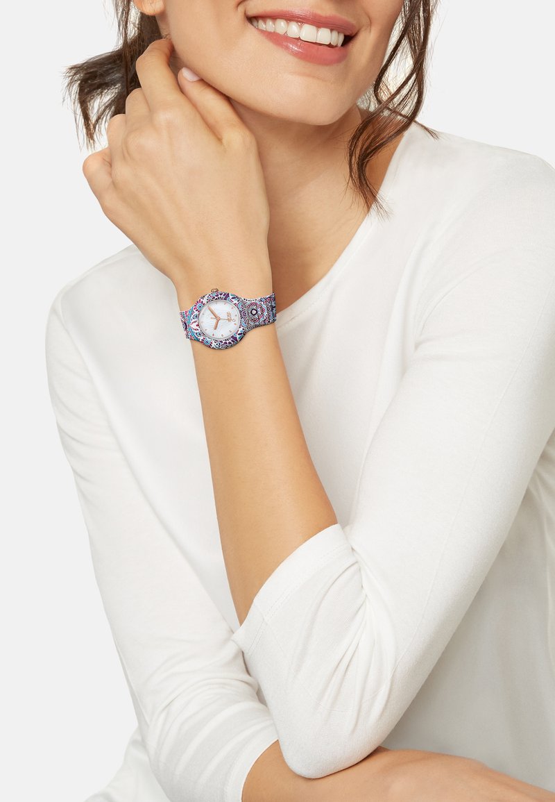 Armbåndsur med en rund hvid urskive, sølvfarvede visere og et farverigt mønstret stofrem med blå, pink og lilla designs.