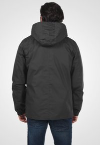 Veste à capuche noire avec des poignets élastiques, texture subtile et coupe décontractée, portée sur un jeans bleu foncé. La vue arrière met en valeur la capuche et les coutures.