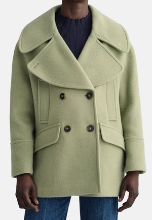 Personne à la peau foncée portant un manteau en laine vert clair à double boutonnage avec de larges revers et des poches à rabat, par-dessus un pull côtelé bleu foncé et un jean.