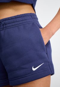 Tamsiai mėlynos medvilninės šortai su elastinga juosta, su šoninėmis kišenėmis ir balta išsiuvinėta Nike logotipu apatiniame krašte.