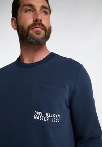 Felpa blu navy in tessuto morbido, con tasca sul petto e dettagli testuali verticali. Collo rotondo e maniche lunghe.