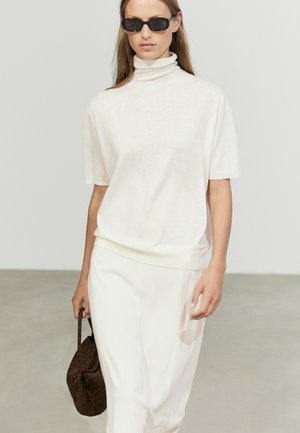 Donna con dolcevita bianco e gonna, che tiene una borsa marrone testurizzata, con capelli lunghi e occhiali da sole scuri.