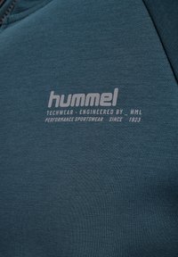 Tessuto techwear in teal con branding integrato, caratterizzato dal logo "hummel", accompagnato da dettagli del prodotto in testo grigio chiaro.