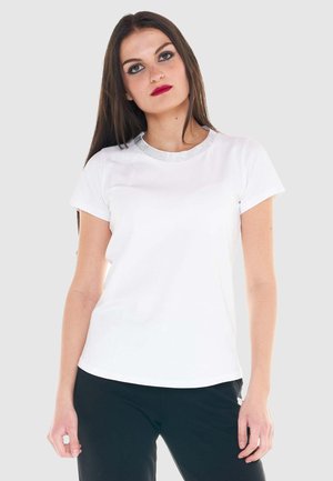 Giovane donna con lunghi capelli castani che indossa una maglietta bianca a maniche corte con scollo decorato e pantaloni neri su sfondo uniforme.