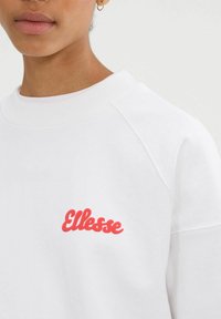 Primer plano de una persona que lleva una sudadera blanca con el logo rojo de "Ellesse" en el pecho y un pequeño pendiente de aro dorado.