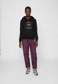 Felpa nera con stampa grafica, abbinata a pantaloni larghi e lucidi di colore viola. Modello che indossa sneakers nere e argentate. Sfondo minimalista.