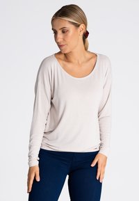 Jasno beżowa bluzka z długim rękawem, szerokim dekoltem i luźnym krojem, wykonana z miękkiego materiału, w zestawieniu z ciemnoniebieskimi dżinsami.