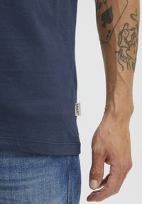 Marineblå bomull T-skjorte med korte ermer, med et lite stoffmerke ved sømmen. Bakgrunnen viser denimjeans.