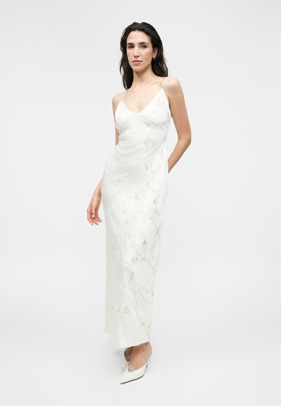 Γυναίκα με μακριά σκούρα μαλλιά, που φοράει λευκό φόρεμα με κεντήματα λουλουδιών και λευκά μυτερά τακούνια, σε απλό φωτεινό φόντο.