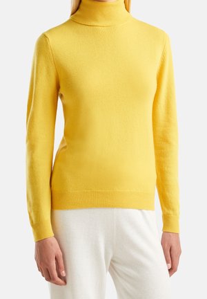 Persona vistiendo un suéter de cuello alto amarillo brillante de manga larga con puños y bajo acanalados, combinado con pantalones blancos, sobre un fondo liso.