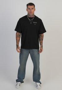 Camiseta negra de gran tamaño con estampado de logo, combinada con jeans de pierna ancha de color azul claro y zapatillas blancas. Presenta brazos tatuados y collares en capas.