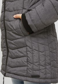 Grå vadderad jacka med dragkedja framtill, muddade ärmslut och en sidoficka. Tyget har en texturerad, melerad yta.