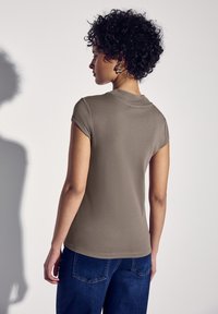 Khaki top s krátkým rukávem a kulatým výstřihem, vyrobený z měkkého jersey materiálu, v kombinaci s modrými denimovými kalhotami, které zdůrazňují přiléhavou siluetu.