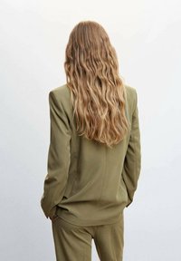 Giacca da abito verde oliva su misura con una vestibilità rilassata, tessuto testurizzato e lunghi capelli ondulati che cadono sulla schiena.