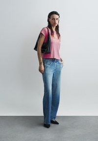 Pink cropped t-shirt with yellow text, high-waisted faded blue jeans, black shoulder bag, and black heels, standing against a plain background.