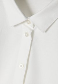 Détail d'un polo blanc présentant un tissu texturé, un col classique et trois boutons en nacre avec un motif floral.