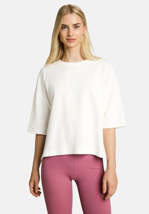 Blonde Frau trägt ein lockeres weißes Kurzarmshirt und eng anliegende pinke Leggings, steht vor einem schlichten hellen Hintergrund.
