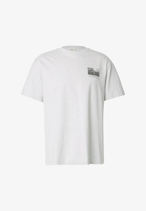 Mężczyzna w jasno szarej koszulce z dwoma czarno-białymi abstrakcyjnymi zdjęciami i małym nadrukiem tekstowym na plecach, stojący na jednolitym tle.