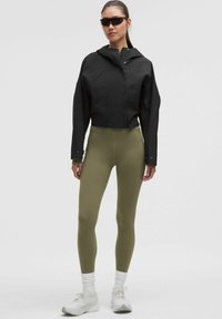 Sort cropped jakke med hætte, grønne leggings og hvide sneakers, med et slankt design og glatte teksturer. Mørke solbriller fuldender looket.