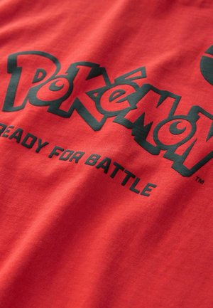 T-shirt en coton rouge avec le texte "Pokémon" en noir et l'impression "PRÊT POUR LE COMBAT". Le tissu a une texture lisse et une coupe décontractée.
