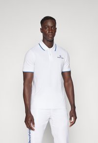 Polo bianco con colletto e polsini blu, con logo sul petto. Indossato con pantaloni bianchi abbinati, che mostrano una vestibilità slim.