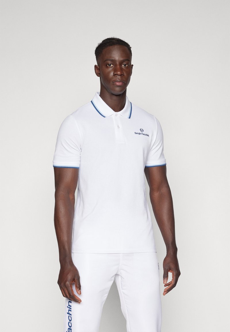 Polo bianco con colletto e polsini blu, con logo sul petto. Indossato con pantaloni bianchi abbinati, che mostrano una vestibilità slim.
