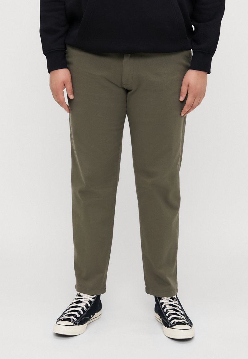 Khaki chinos sirge lõikega, koos mustade kõrge säärega tossudega. Materjal on sile ja klassikalise nelja taskuga disainiga.