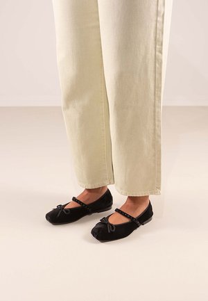 Bailarinas de ante negro con punta redondeada, lazo decorativo y tira con cuentas, combinadas con pantalones de bota ancha de color claro.