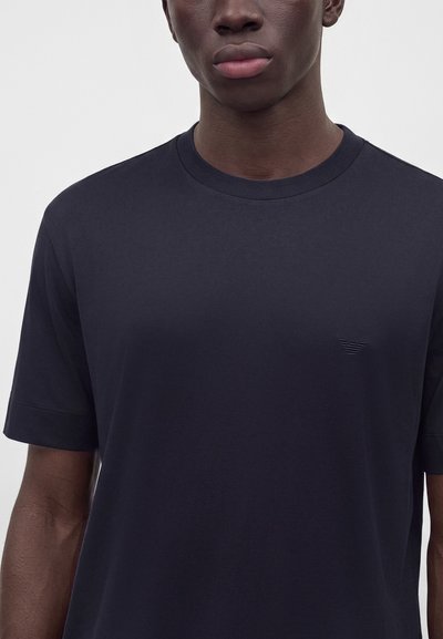 Νέος που φοράει ένα απλό σκούρο μπλε T-shirt με στρογγυλή λαιμόκοψη και μικρό ανάγλυφο λογότυπο στο αριστερό μέρος του στήθους.