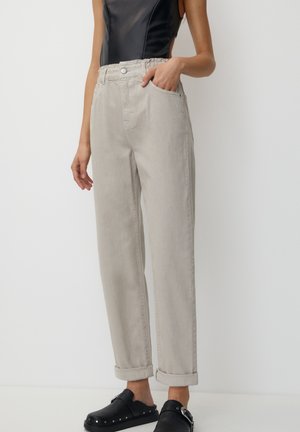 Donna che indossa pantaloni beige a vita alta con risvolti, top senza maniche nero e scarpe scivolate nere con borchie e fibbie argentate, in piedi contro un muro bianco.