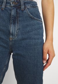 Korkeavyötäröiset tummansiniset farkut, joissa on sileä rakenne, oranssit ompeleet ja etutasku. Käsivarsi lepää sivulla.