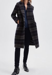Plaid wool coat in dark gray and navy, featuring two front pockets, double-breasted buttons, and a relaxed fit. Paired with dark pants and boots.
